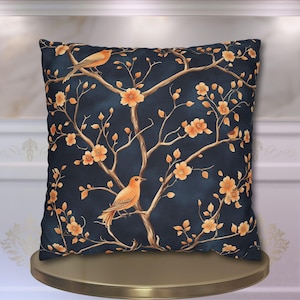 May include: Square decorative pillow with a dark blue background featuring a tree branch design with orange flowers and birds. The pillow is placed on a gold-colored stand.