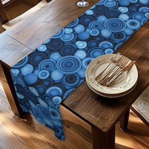 May include: A blue table runner with a repeating pattern of concentric circles in varying shades of blue. The runner is placed on a wooden table, with a set of plates, forks, and knives visible on the right side.