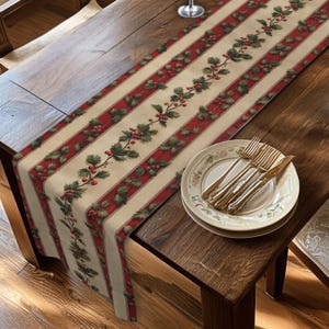 May include: A long rectangular table runner featuring a cream-colored background with vertical red stripes and a repeating pattern of green holly leaves and red berries. A stack of two white plates with gold rims and gold cutlery sits on top.