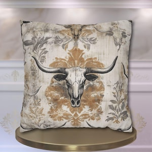 May include: A decorative throw pillow with a white background and a gold and grey damask pattern. A black and white illustration of a bull skull is centred on the pillow.