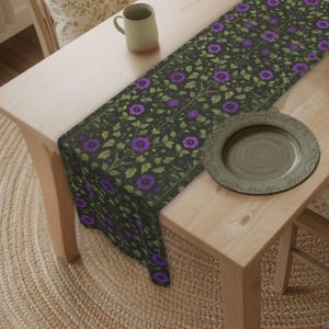 May include: A rectangular table runner featuring a dark charcoal background with a repeating pattern of vibrant purple flowers and light green foliage. The runner is draped across a light-toned wooden table, accompanied by a ceramic mug and a plate.