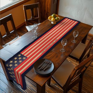 May include: A patriotic table runner featuring the American flag design. The runner has red and beige stripes with a blue section displaying gold stars. Text reads "250th Anniversary 1776-2026".