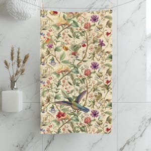 May include: A cream-colored bath towel with a floral and hummingbird design. The towel features a repeating pattern of flowers in shades of pink, purple, and yellow, with green leaves and vines. A hummingbird with blue and green wings is in flight.