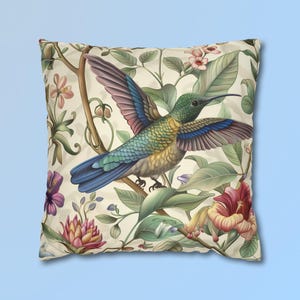 May include: A square throw pillow featuring a central hummingbird with iridescent green, blue, and brown feathers in flight. The background displays a botanical pattern of light cream fabric with illustrations of pink, purple, and red flowers.