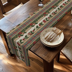 May include: A rectangular table runner featuring a festive pattern of green holly leaves and red berries arranged in vertical stripes, interspersed with thin red and green plaid bands on a cream-colored background, draped over a wooden table.