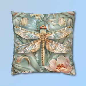 May include: A square throw pillow featuring a central dragonfly with translucent, veined wings and a tan body. The background displays a soft green botanical pattern with pale pink and cream-colored flower blossoms surrounding the insect.