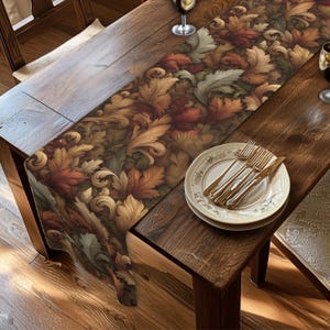 May include: A long rectangular table runner featuring a dense, repeating pattern of autumn-colored leaves in shades of deep red, burnt orange, golden yellow, and muted green, draped across a dark wooden table with a place setting of plates and cutlery.