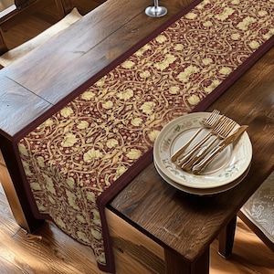 May include: A decorative table runner featuring a burgundy and gold floral design. The runner is placed on a dark wooden table. A set of plates, forks, and knives are on the table, adding to the dining setting.