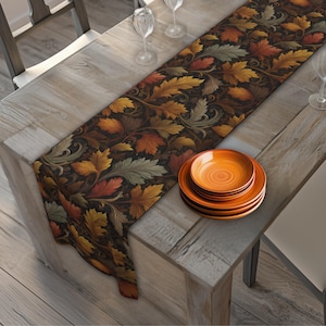 May include: A long table runner featuring a dark brown background with a repeating pattern of autumn leaves in shades of orange, yellow, and muted green. A stack of three round orange plates sits on the runner, alongside two clear glass goblets.