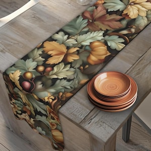 May include: A table runner with a botanical design featuring autumn leaves in shades of green, brown, and gold. A stack of four brown ceramic plates sits on a wooden table, complementing the runner's colors. The table is made of light-colored wood.