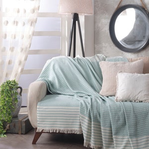 May include: A light blue and white striped throw blanket with fringe edges draped over a white couch. The blanket has a chevron pattern and is made of a soft, textured fabric.