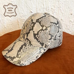 Pode incluir: Um boné de beisebol com um padrão de pele de cobra cinzento e branco e um acabamento texturizado. O boné inclui uma etiqueta no canto superior esquerdo com o texto Genuine 100% Leather. O design consiste numa copa arredondada e aba curva.