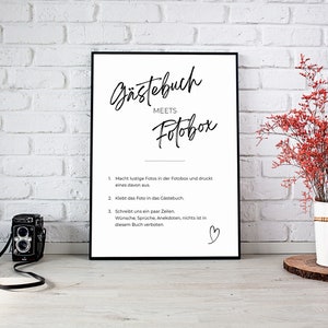 Puede incluir: Una impresión en blanco y negro enmarcada con el texto "Gästebuch MEETS Fotobox" e instrucciones. Una cámara vintage está frente a la impresión. Un jarrón de flores rojas está a la derecha.