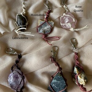 May include: Assortment of crystal keychains, including Labradorite, Moonstone, Rose Quartz, Amethyst, and Moss Agate. Each crystal is wrapped in a macrame design and attached to a silver-colored clasp. The names of the crystals are labeled in the image.
