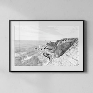 May include: A black and white landscape photograph in a thin black frame. The image depicts a rugged coastline with rocky cliffs overlooking the ocean, featuring crashing waves and a distant horizon line under a bright, clear sky.