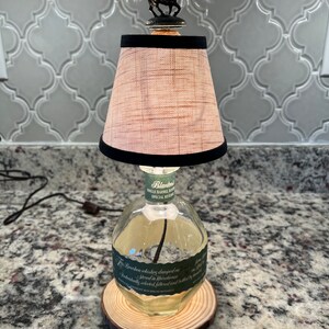 May include: A repurposed Blanton's Single Barrel Bourbon bottle lamp with a brown and black lampshade and a jockey riding a horse figurine on top. The bottle is clear and has a green label with the text "Blanton's Single Barrel Bourbon Special Reserve" and "The bourbon whiskey dumped on stored in warehouses...", and is sitting on a wooden base.