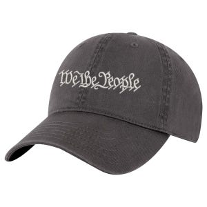 May include: A charcoal gray baseball cap featuring the text We the People embroidered in white thread across the front panel. The cap has a curved brim, a six-panel construction with stitched eyelets, and a structured crown with a button on top.