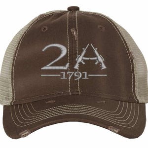 May include: A brown distressed trucker cap featuring a mesh back and a solid front panel. The front is embroidered with the silver text 2A, a graphic of two crossed rifles, and the year 1791 centered below the rifles with a horizontal line.
