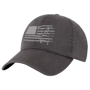 Peut inclure: Une casquette de baseball gris anthracite avec un motif brodé sur le panneau avant. La broderie représente un drapeau américain dont les rayures sont composées de diverses silhouettes de fusils, réalisées en fil gris clair.