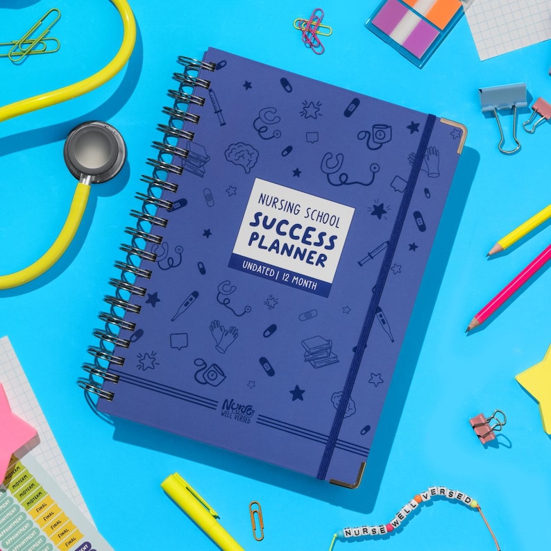 May include: A spiral-bound, navy blue Nursing School Success Planner with the text "Nursing School Success Planner Undated | 12 Month". The planner is decorated with medical-themed icons and a matching elastic band. A yellow stethoscope and other stationery items surround the planner.