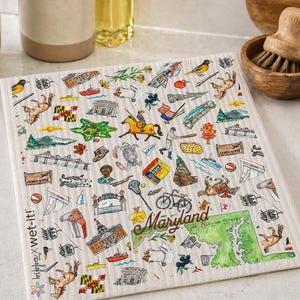 May include: A square, off-white dishcloth with a Maryland-themed design. The cloth features colorful illustrations of landmarks, animals, and symbols of Maryland. The word "Maryland" is written in a cursive font. A wooden bowl with a brush is next to the cloth.