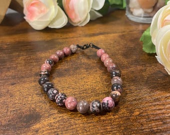 May include: A beaded bracelet featuring a mix of pink, black, and gray stones. The bracelet is strung on a black cord and is displayed on a wooden surface. The stones vary in size and pattern, creating a unique accessory.