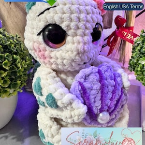 May include: A white crocheted plush creature with large dark eyes, pink cheeks, and teal spots, holding a purple crocheted shell with a white pearl bead. The image includes text labels for PDF file download and English USA terms.