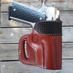 May include: A brown leather holster with white stitching for a handgun. The holster has a belt loop and a cut-out for a thumb break. The holster is designed to be worn on the hip.