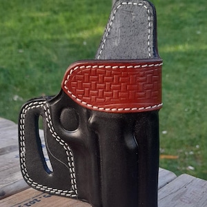 May include: A black leather holster with white stitching. The holster features a brown, textured band with white stitching. The holster is designed to be worn on the hip. The holster is made of leather.