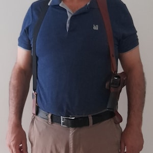 May include: A person wearing a blue polo shirt with a brown leather shoulder holster and a black belt. The holster is holding a black handgun.