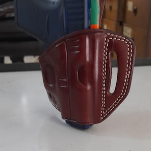 Könnte beinhalten: Ein braunes Lederholster mit weißen Nähten für eine Pistole. Das Holster hat eine Gürtelschlaufe und einen Abzugsschutz.