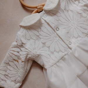 K&ouml;nnte beinhalten: Wei&szlig;es Babykleid mit floraler Spitzenborte und T&uuml;llrock. Das Kleid hat lange &Auml;rmel und einen Bubikragen. Das Kleid h&auml;ngt an einem Holzkleiderb&uuml;gel.
