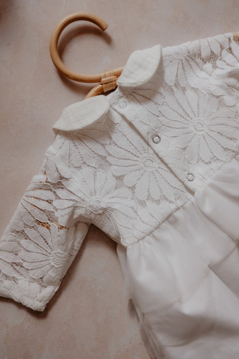 K&ouml;nnte beinhalten: Wei&szlig;es Babykleid mit floraler Spitzenborte und T&uuml;llrock. Das Kleid hat lange &Auml;rmel und einen Bubikragen. Das Kleid h&auml;ngt an einem Holzkleiderb&uuml;gel.