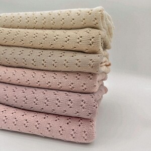 May include: A stack of six folded blankets in varying shades of beige and pink. The blankets have a delicate, openwork pattern. The colors range from a light cream to a deep rose, creating a soft, inviting aesthetic. The blankets are neatly stacked.
