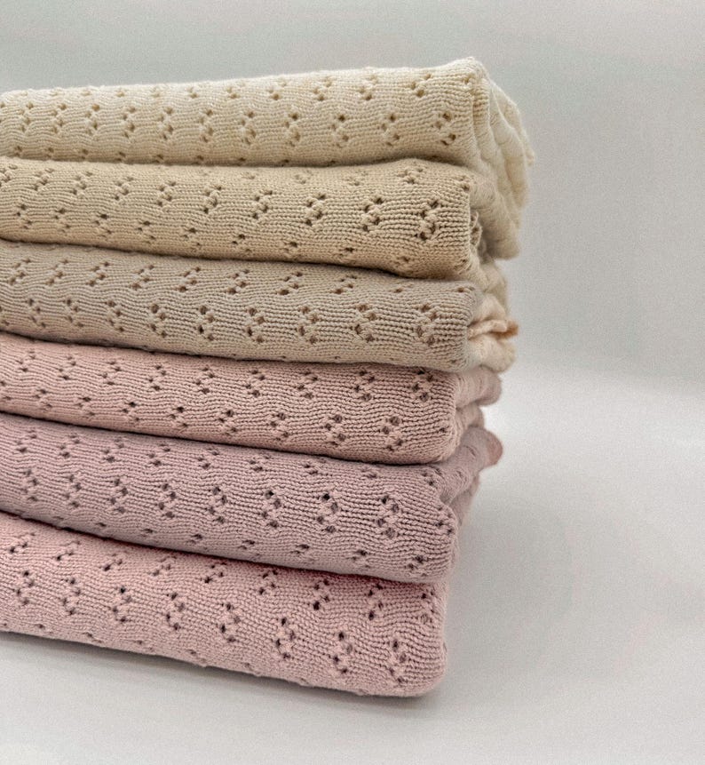 May include: A stack of six folded blankets in varying shades of beige and pink. The blankets have a delicate, openwork pattern. The colors range from a light cream to a deep rose, creating a soft, inviting aesthetic. The blankets are neatly stacked.