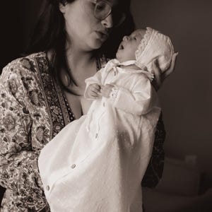 以下が含まれることがあります： セピアトーンの画像。白いガウンとボンネットを着た赤ちゃんが、メガネと柄物のローブを着た人に抱かれています。赤ちゃんのガウンはテクスチャパターンとレースのトリムが付いています。ボンネットにもレースのトリムが付いています。