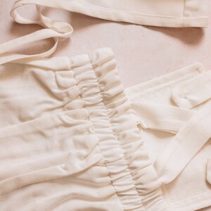 May include: Cream-colored baby clothing set, including a bonnet and pants with an elastic waistband. The fabric appears to be soft and lightweight, suitable for a newborn. The set is laid out on a light pink surface.