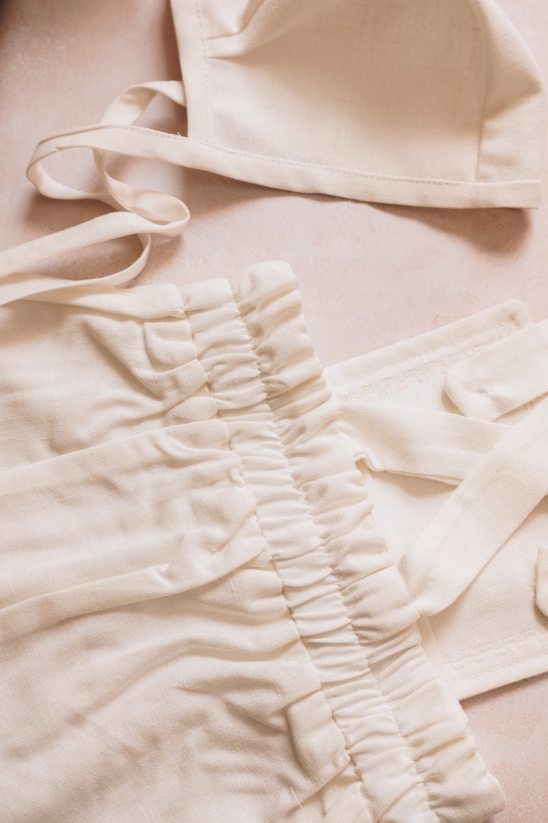 May include: Cream-colored baby clothing set, including a bonnet and pants with an elastic waistband. The fabric appears to be soft and lightweight, suitable for a newborn. The set is laid out on a light pink surface.