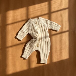May include: Cream-colored baby romper with a ribbed knit design. The one-piece garment features long sleeves, a button-up front, and wooden buttons. The romper is made of a soft, textured material and is designed for comfort and style.
