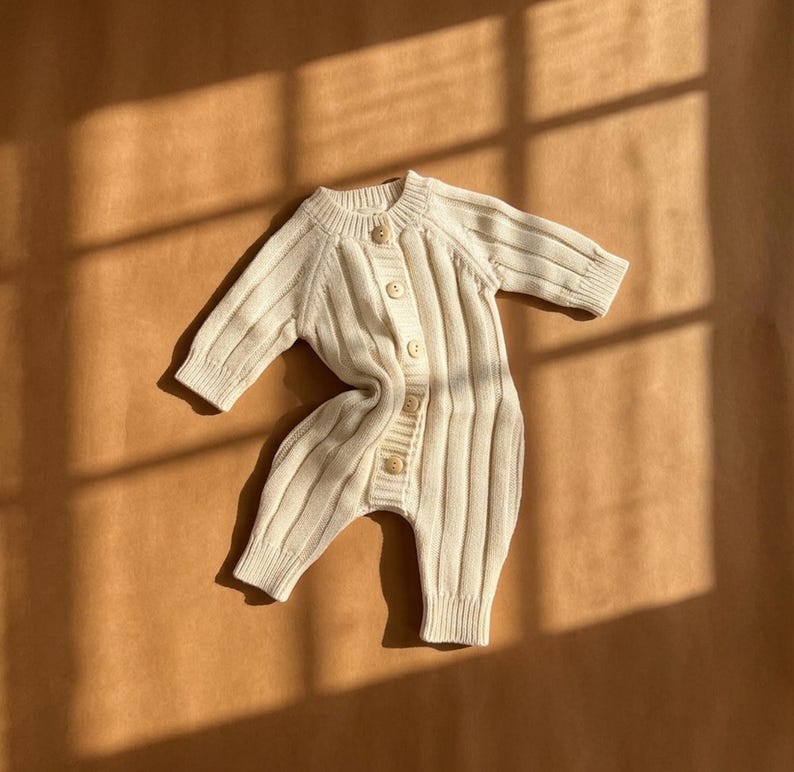May include: Cream-colored baby romper with a ribbed knit design. The one-piece garment features long sleeves, a button-up front, and wooden buttons. The romper is made of a soft, textured material and is designed for comfort and style.