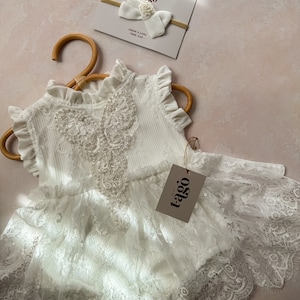 May include: A white lace baby romper with a ruffled neckline and sleeves, hanging on a wooden hanger. A matching white bow headband with pearl accents and a tag with the text "tago" are also visible.