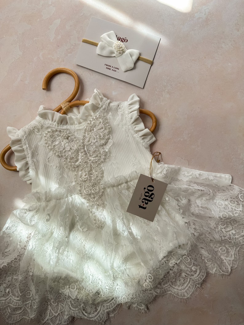 May include: A white lace baby romper with a ruffled neckline and sleeves, hanging on a wooden hanger. A matching white bow headband with pearl accents and a tag with the text "tago" are also visible.