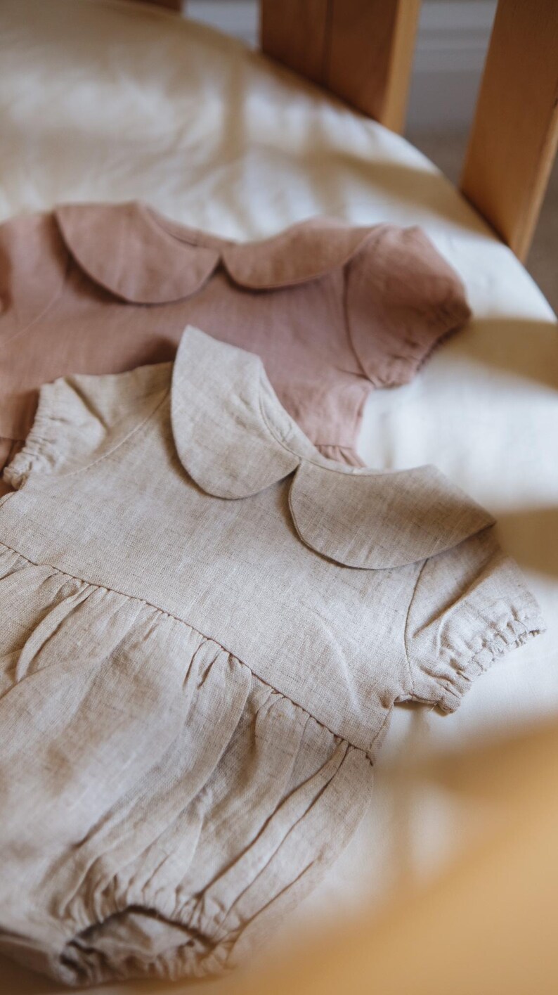 Puede incluir: Dos peleles de beb&eacute; de lino en colores neutros suaves. Uno es beige claro, el otro rosa apagado. Ambos tienen cuello Peter Pan y mangas cortas. La tela parece ligera y transpirable, ideal para el verano.