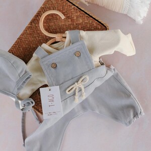Op de afbeelding: Een lichtblauwe baby-outfitset, bestaande uit een tuinbroek, een shirt met lange mouwen en een muts. De tuinbroek heeft houten knopen en een decoratieve strik. De set wordt gepresenteerd met een rieten mand en een cr&egrave;mekleurige deken.