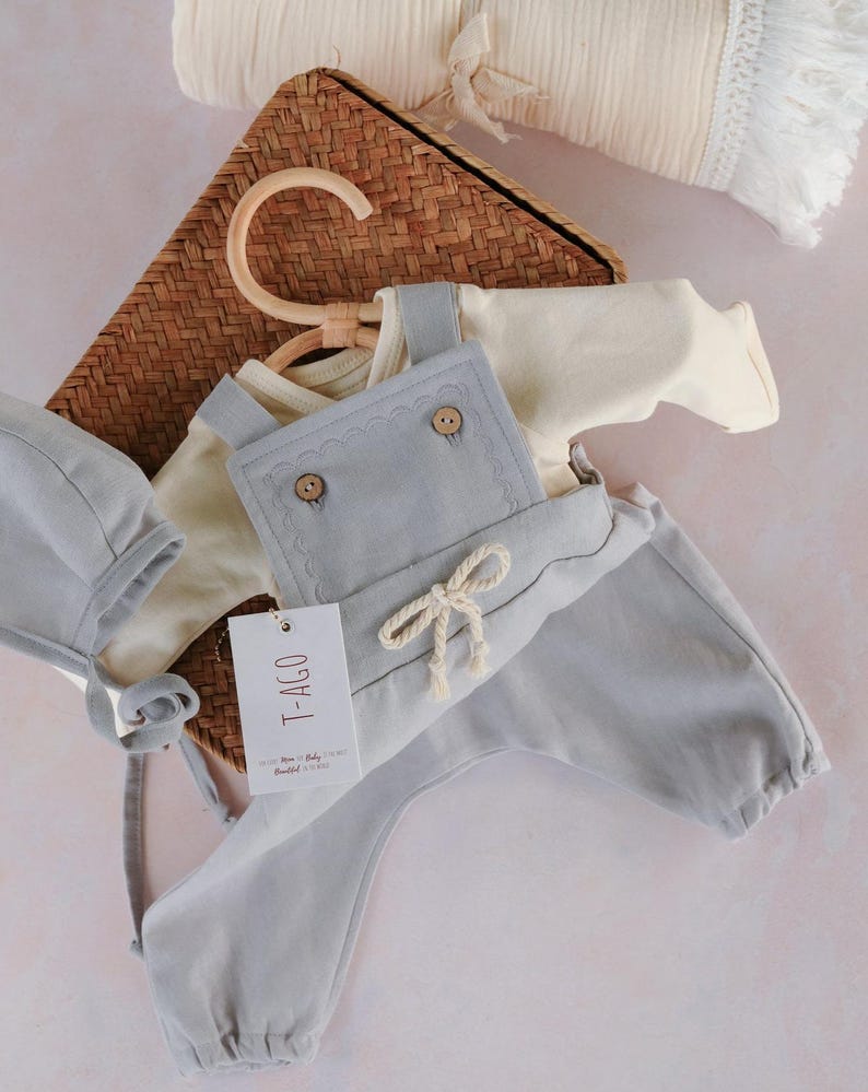 Op de afbeelding: Een lichtblauwe baby-outfitset, bestaande uit een tuinbroek, een shirt met lange mouwen en een muts. De tuinbroek heeft houten knopen en een decoratieve strik. De set wordt gepresenteerd met een rieten mand en een cr&egrave;mekleurige deken.