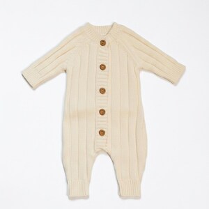 May include: Cream-colored baby romper with long sleeves and ribbed knit texture. The one-piece garment features a button-up front with six wooden buttons. The romper has a round neckline and ribbed cuffs and ankles.