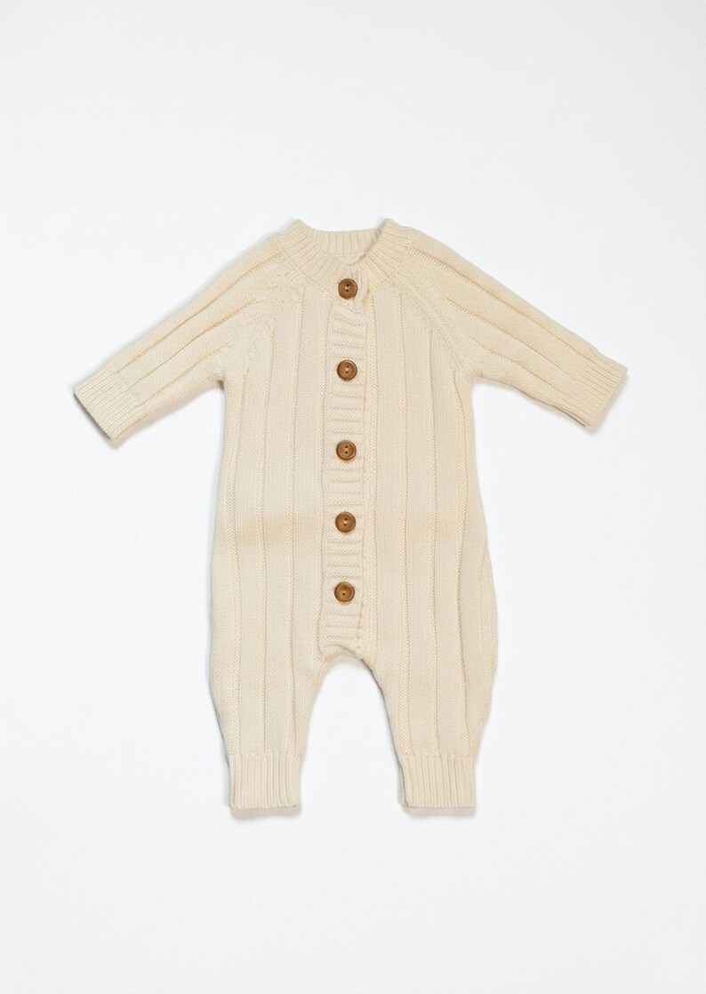 May include: Cream-colored baby romper with long sleeves and ribbed knit texture. The one-piece garment features a button-up front with six wooden buttons. The romper has a round neckline and ribbed cuffs and ankles.