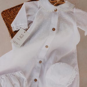 以下が含まれることがあります： 襟付きシャツ、ブルマ、ボンネットを含む白いベビー服セット。木製のボタンとレースのトリムが特徴です。「T-AGO」と書かれたタグが付いています。柳のバスケットと木製のハンガーにセットが展示されています。