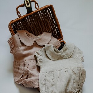 Puede incluir: Dos peleles para beb&eacute;s en colores suaves y apagados, uno rosa empolvado y otro beige claro, se exhiben cerca de una cesta de mimbre. Cada pelele presenta un cuello Peter Pan y detalles fruncidos, lo que sugiere un estilo vintage o cl&aacute;sico. La tela parece ser lino o un material ligero similar.