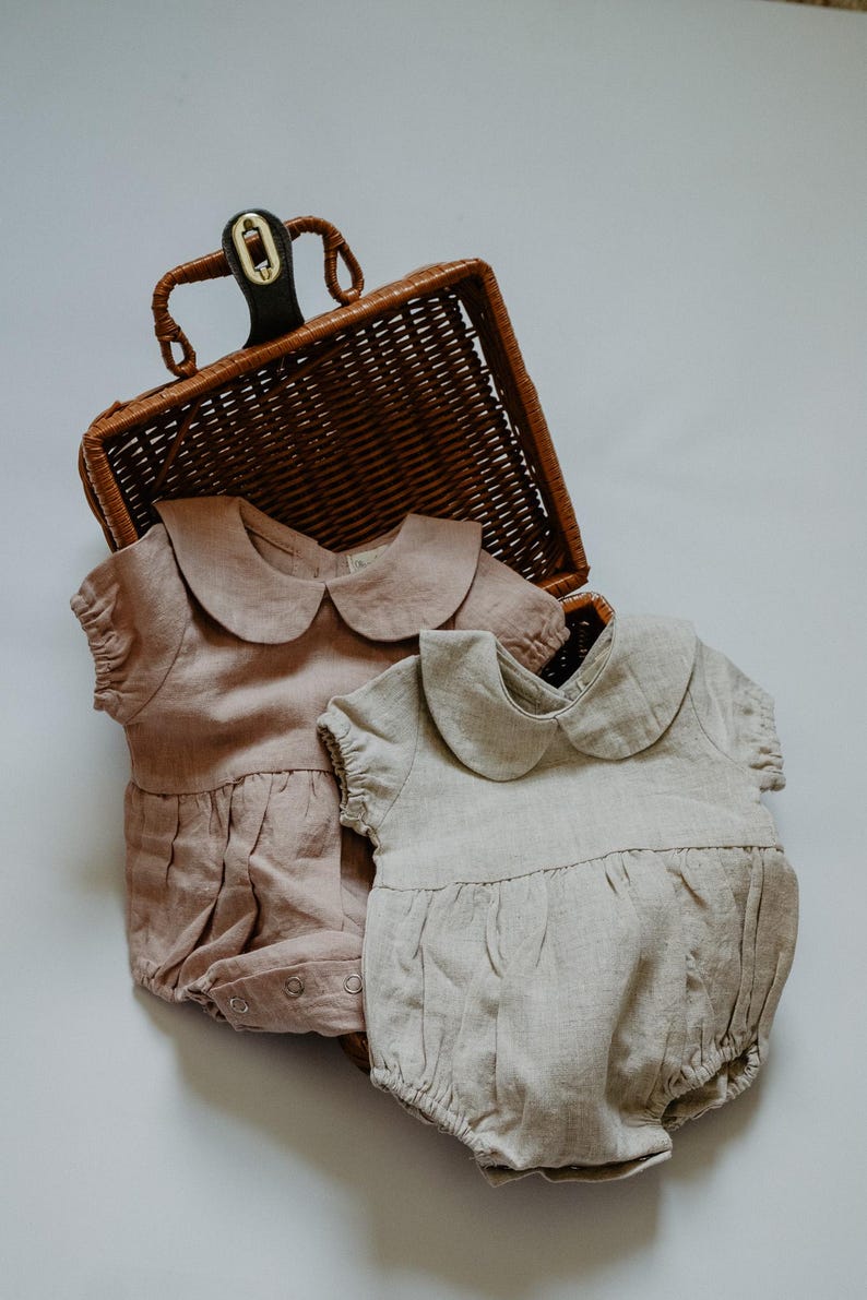 Puede incluir: Dos peleles para beb&eacute;s en colores suaves y apagados, uno rosa empolvado y otro beige claro, se exhiben cerca de una cesta de mimbre. Cada pelele presenta un cuello Peter Pan y detalles fruncidos, lo que sugiere un estilo vintage o cl&aacute;sico. La tela parece ser lino o un material ligero similar.